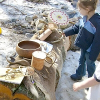 070303WampanoagSpoonEtAl1 The exhibit log holds a furry deer hide, a wooden bowl and birch-bark baskets, a deerskin drum, and more for kids to touch and feel. In front on the notebook, a toy made of a deer's leg bone and some of its (hollow) toe bones, strung together with a deer-hide thong. The game is to hold the leg bone sharp end up, and use it to swing the toe bones into the air. It's not easy to skewer one of them on the leg bone!  Just behind this toy, is an strange-looking piece of dark wood which is...